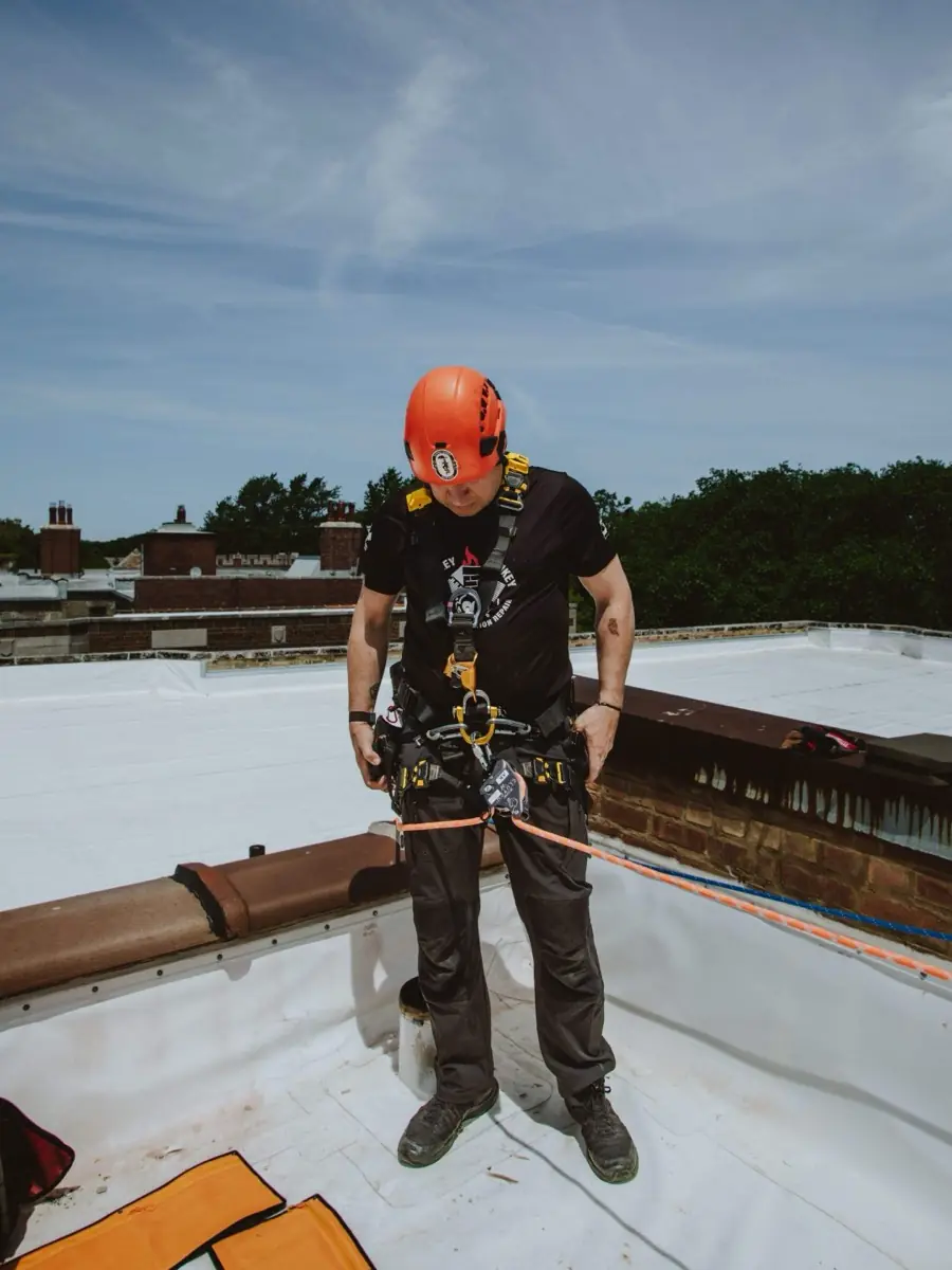 Rope access technician in a red helmet and full harness standing on a white flat membrane roof in Chicago, checking equipment with ropes running toward the parapet