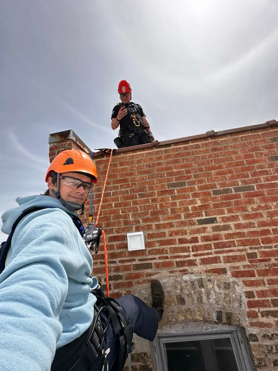 Two rope access technicians in orange and red helmets and harnesses at the brick parapet of a Chicago rooftop, with the dryer vent cover visible on the wall below them