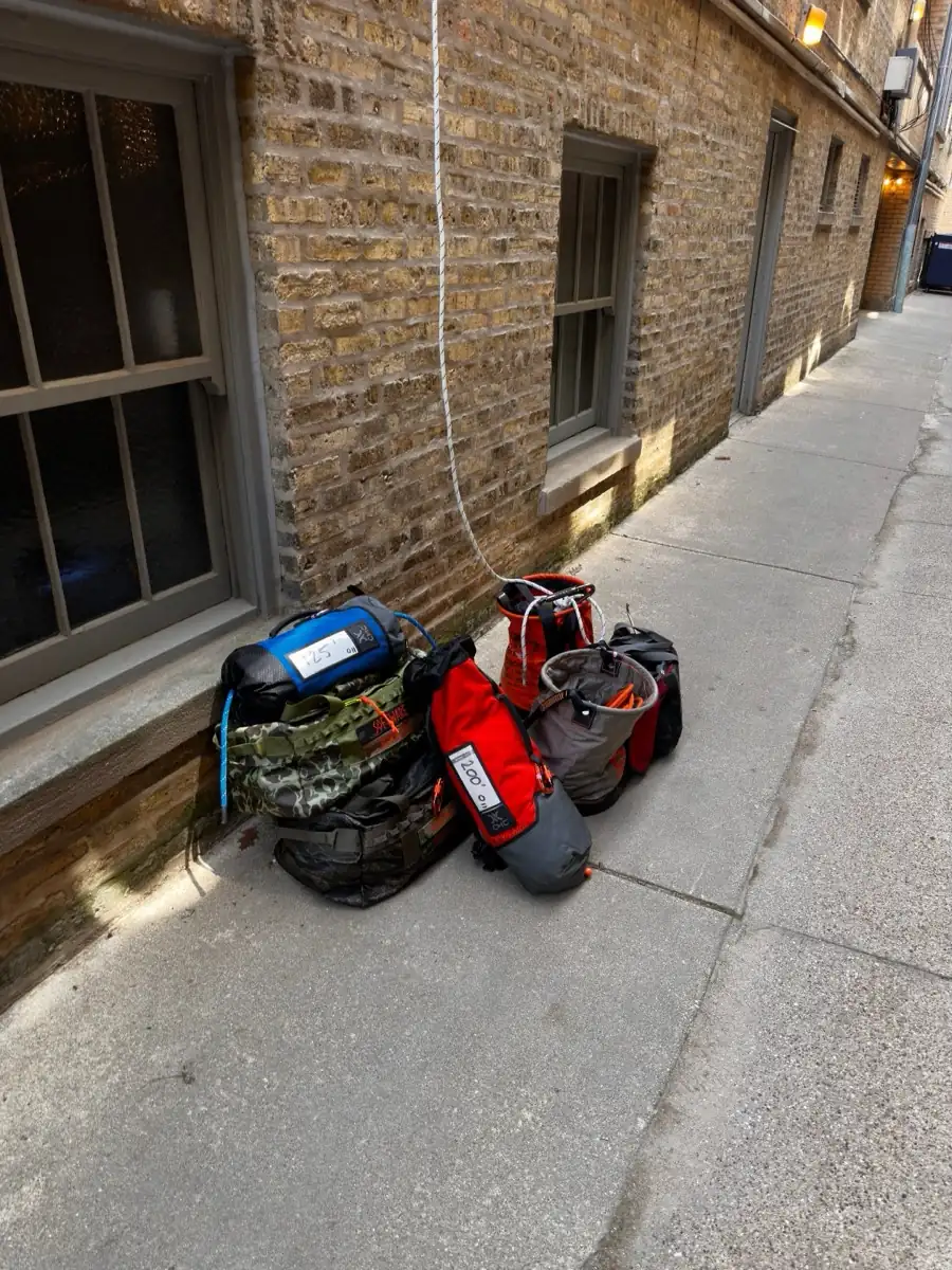 Rope access equipment bags staged on the concrete of a narrow Chicago gangway beside a brick wall, with a rope hanging down from the roof above