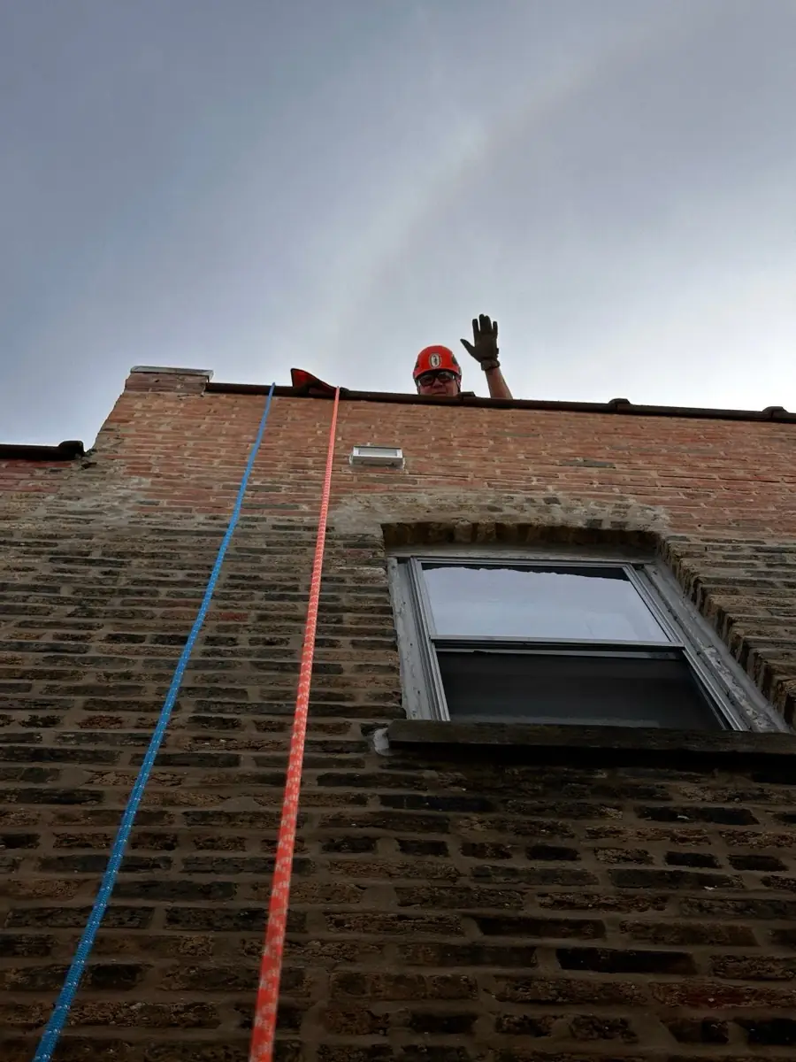 View looking up a Chicago brick wall to the roofline, showing the louvered metal dryer vent cover in place and a technician in an orange helmet at the parapet with two ropes running down the wall