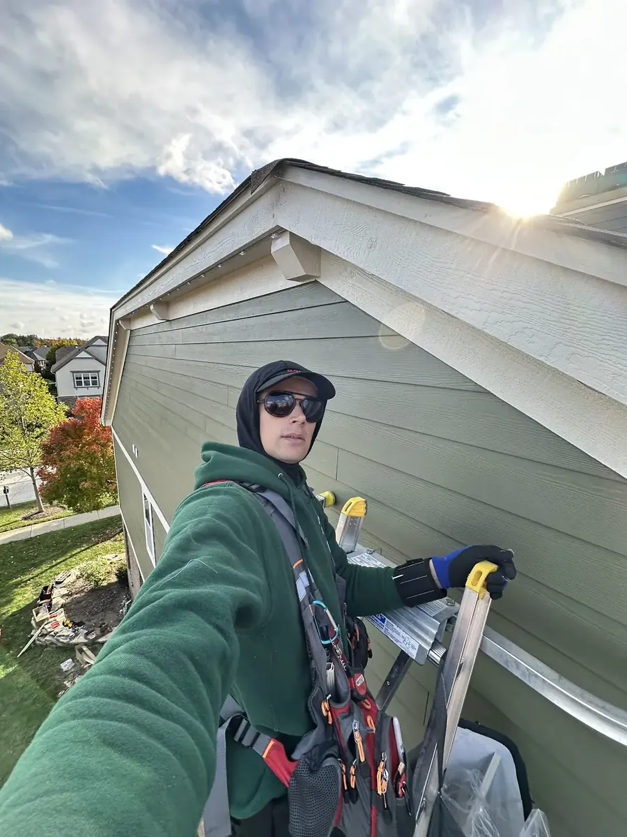 Hanging Lights Club installer in a safety harness on a ladder at the roofline of a two-story home, fitting permanent lighting track under the eave