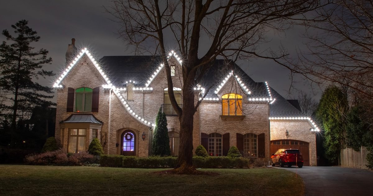 We Clean Your Roofline All Year. Hanging Lights Club Lights It, Halloween Through Valentine's Day Brick home outlined in warm white roofline lighting at dusk with bare winter trees in the front yard