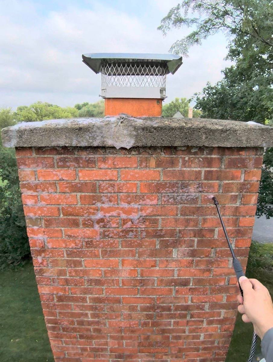 Brick chimney above the roofline being rinsed with low pressure water from a wand after roof soft washing, with the metal chimney cap visible on top