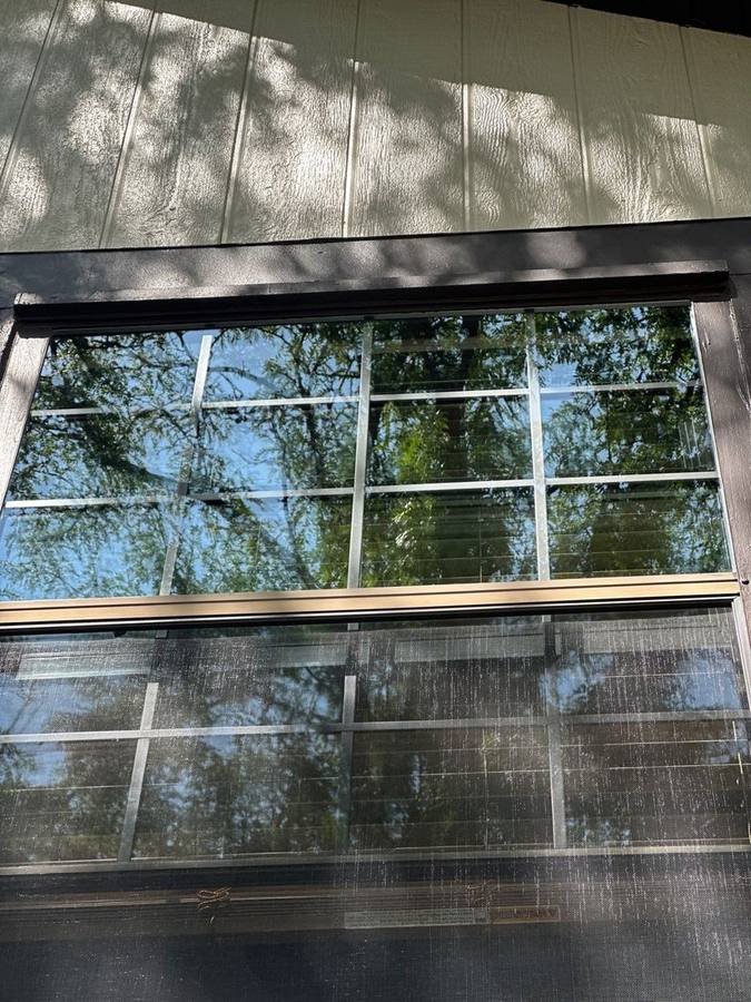 A single window photographed after cleaning, with the upper sash glass clear enough to show sharp tree branches and blue sky, and the lower half still behind a screen where the same view appears grey and hazy