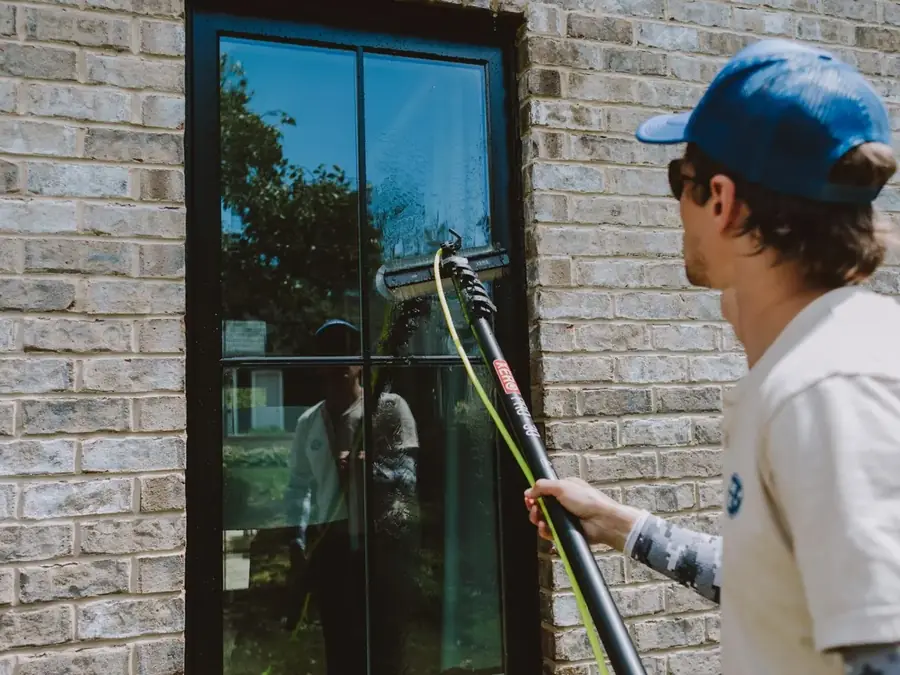 Professional window cleaner using long-reach pole to clean exterior windows on brick home