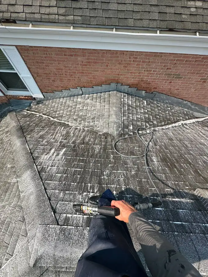 Pane Pal technician on an Inverness roof applying soft wash solution through a low pressure wand, hose run over the ridge, brick chimney wall and roof valley visible below